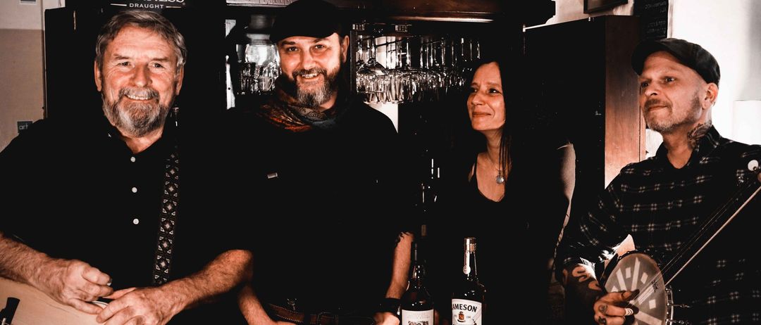 Vier Personen stehen lächelnd um ein Fass in einer Bar. Zwei Whiskeyflaschen stehen auf dem Fass. Dahinter befindet sich ein Regal mit Gläsern und einem Guinness-Schild.