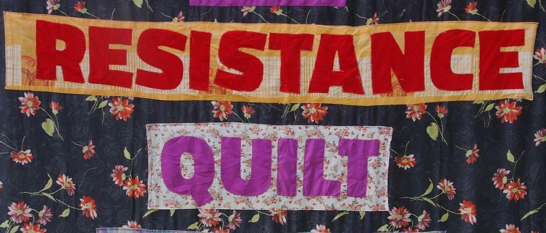 Eine farbenfrohe Decke mit 'The Resistance Quilt Project', umgeben von Blumen- und Punktemustern im Hintergrund.