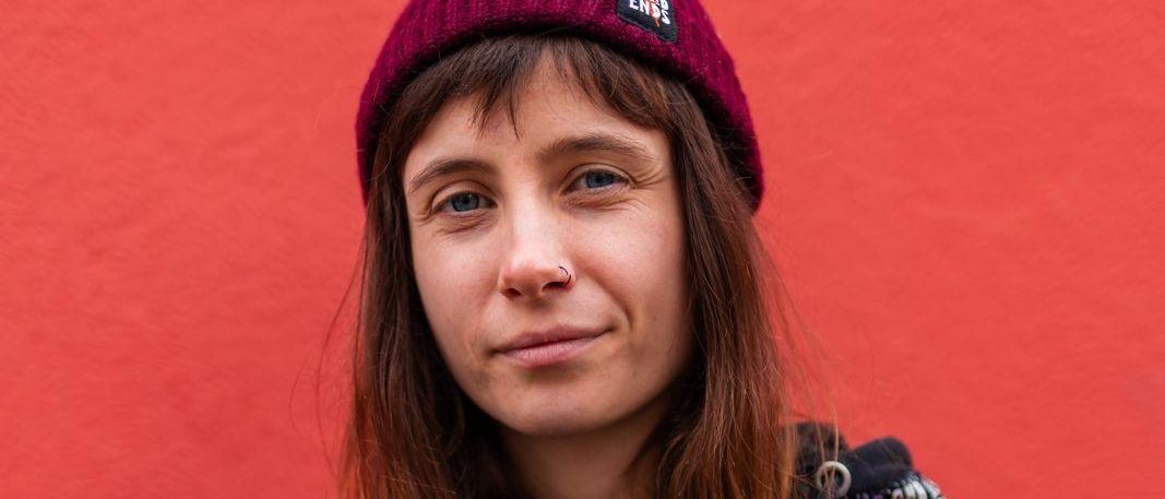 Eine Frau mit langen braunen Haaren, die eine lila Leopardenmuster-Jacke und eine rote Mütze trägt, steht vor einer roten Wand.