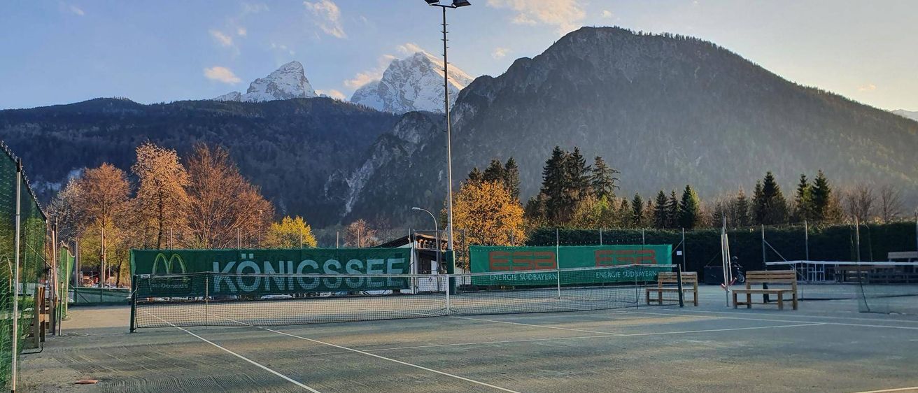Ein Tennisplatz mit einem Netz vor einem Schild mit grünem Text auf einem Banner. Der Platz ist von Bäumen und Bergen umgeben.