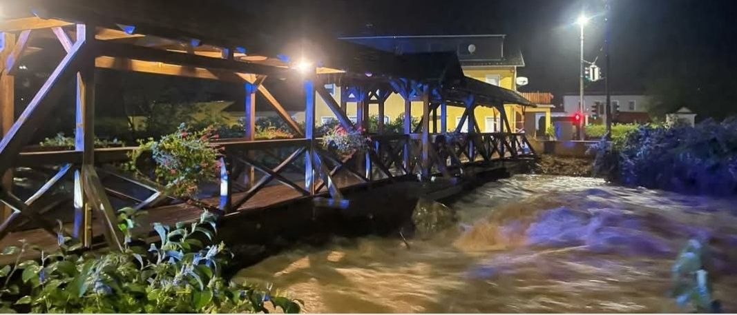 Nachtaufnahme einer Brücke über einem überfluteten Bereich. Die Brücke ist mit blauen Lichtern beleuchtet. Der Textüberlagerung lautet 'Infoveranstaltung: Starkregen, Hochwasser und Selbstschutz.' Datum ist der 27. August.