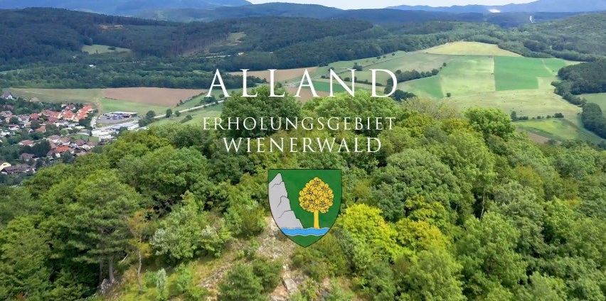 Ein Luftbild von Alland, einem Erholungsgebiet mit üppigen grünen Bäumen, Bergen und einem Dorf darunter. Das Bild zeigt das Logo der Region.