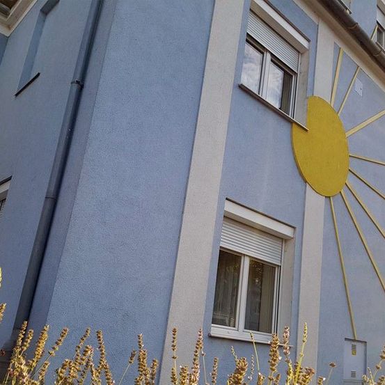 Ein blaues Gebäude mit einem gelben Sonnendesign an der Wand, mit drei Fenstern mit weißen Rollläden und einem kleinen Garten mit Lavendelblumen davor.