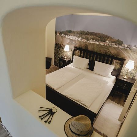 Ein Schlafzimmer mit einem Doppelbett, weißen Bettlaken und zwei Nachttischen mit Lampen. Ein Hut und Schlüssel liegen auf der Kopfseite des Bettes. Die Wand ist mit einer landschaftlichen Tapete verziert.