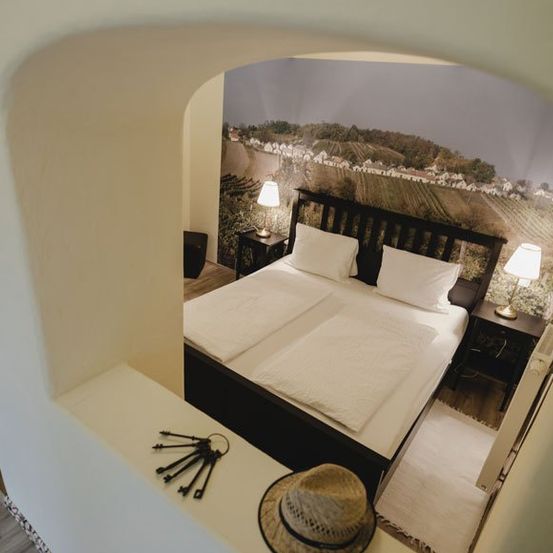 Ein Schlafzimmer mit einem Doppelbett, weißen Bettlaken und zwei Nachttischen mit Lampen. Ein Hut und Schlüssel liegen auf der Kopfseite des Bettes. Die Wand ist mit einer landschaftlichen Tapete verziert.