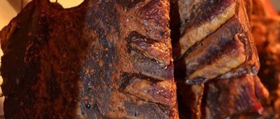 A close-up view of a well-cooked piece of meat, possibly ribs, hanging in a row with others. The meat has a charred and smoky appearance.