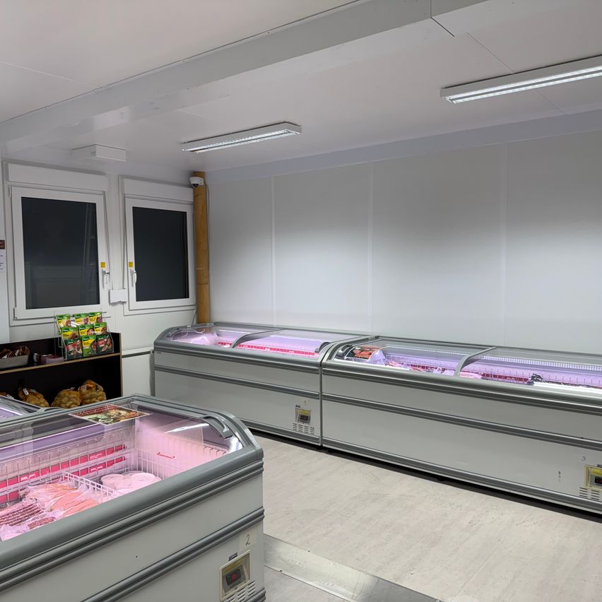 Innenansicht eines Kühllagers. Es enthält mehrere Gefrierschränke mit Fleischprodukten, weiße Wände, Decke mit Leuchtstofflampen und einen Regal mit einigen Produkten auf der linken Seite.