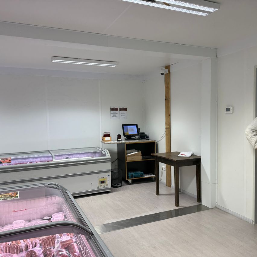 Ein leerer Ladeninnenraum mit zwei Kühlvitrinen für Fleisch. Ein Schreibtisch mit Computermonitor und Regal steht an der Wand. Es gibt Leuchten an der Decke und eine Überwachungskamera.