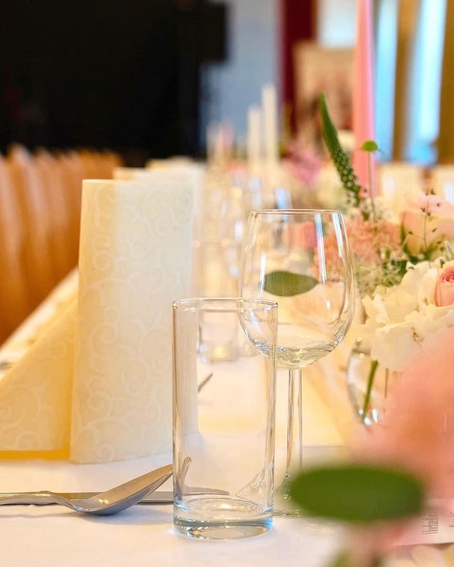 Ein Tisch für eine formelle Veranstaltung mit einem Weinglas, einem kleinen Glas und Blumen im Hintergrund. Es gibt zwei gemusterte Servietten und der Tisch ist mit einer weißen Tischdecke bedeckt.