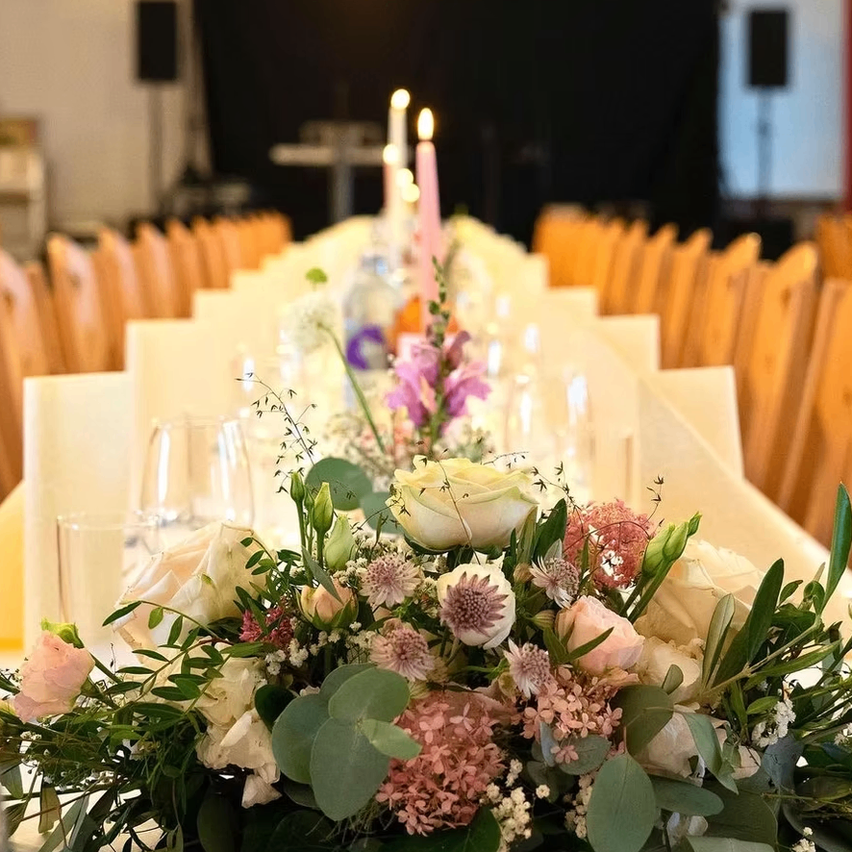 Ein langer Tisch mit Blumenarrangements, Weingläsern und brennenden Kerzen in einem Bankettsaal.