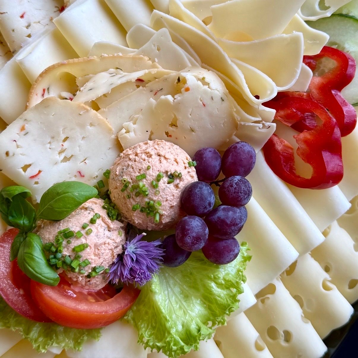 A plate with various cheeses, grapes, tomatoes, red peppers, and herbs. Two cheese balls are topped with green onions and purple flowers.