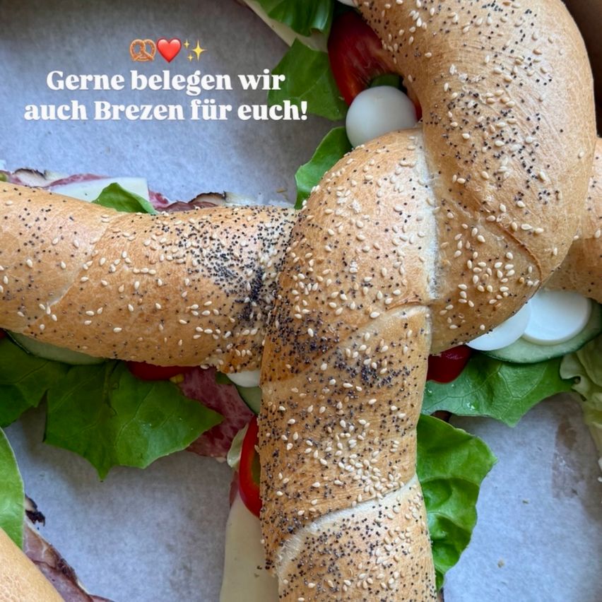 A close-up of a pretzel sandwich with lettuce, tomato, and cheese, displayed on a parchment-covered surface. German text overlay reads 'Gerne belegen wir auch Brezen für euch!'.