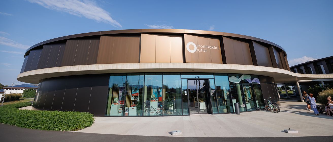 Das Shoemakers Outlet Gebäude zeichnet sich durch eine geschwungene Fassade mit braunen Paneelen und großen Glasfenstern aus. Der Eingang hat eine Glastür und einen Mülleimer. Im Inneren sind verschiedene Artikel ausgestellt.