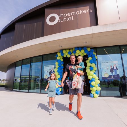 Ein Mann und zwei Kinder gehen Hand in Hand vor einem Shoemakers Outlet-Geschäft. Der Eingang ist mit bunten Luftballons geschmückt.