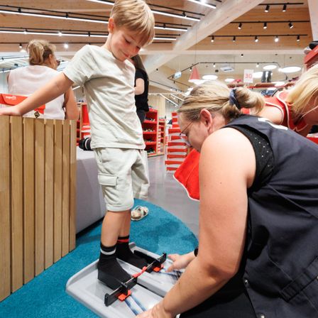 Ein kleiner Junge steht auf einer Messwaage, während eine Frau seine Fußgröße in einem Schuhgeschäft überprüft. Eine andere Frau beobachtet im Hintergrund.