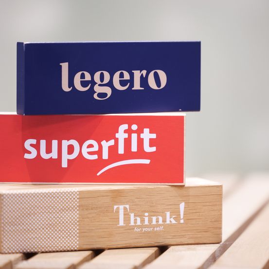 Drei Holzkisten, die aufeinandergestapelt sind. Die oberste ist blau mit weißem Text 'legero'. Darunter ist eine rote Box mit weißem Text 'superfit'. Die unterste hat weiße Punkte und den Text 'Denke!'