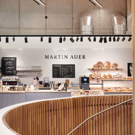 Eine Bäckerei mit einer geschwungenen Holztheke. Der Name 'Martin Auer' ist an der Wand angebracht. Es gibt Tafeln mit Text, Regale mit Brot und verschiedene Gegenstände auf der Theke.