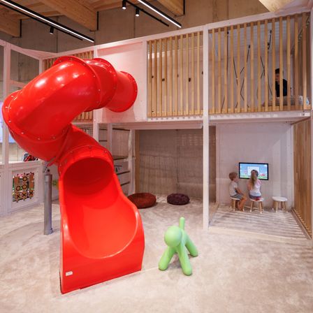 Ein Indoor-Spielbereich mit einer roten Spiralrutsche, einem Holzbalkon und einem Monitor. Zwei Kinder schauen fern, während eine Person auf dem Balkon steht.