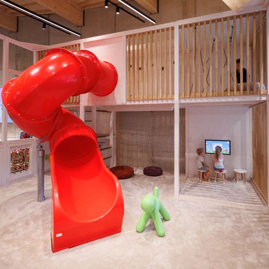 Ein Indoor-Spielbereich mit einer roten Spiralrutsche, einem Holzbalkon und einem Monitor. Zwei Kinder schauen fern, während eine Person auf dem Balkon steht.
