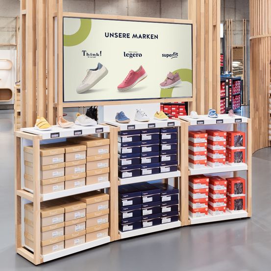 Ein Verkaufsdisplay mit Regalen voller Schuhkartons. Ein großer Bildschirm darüber zeigt die Marken 'Think!', 'Legero' und 'Superfit' mit Bildern von Schuhen.