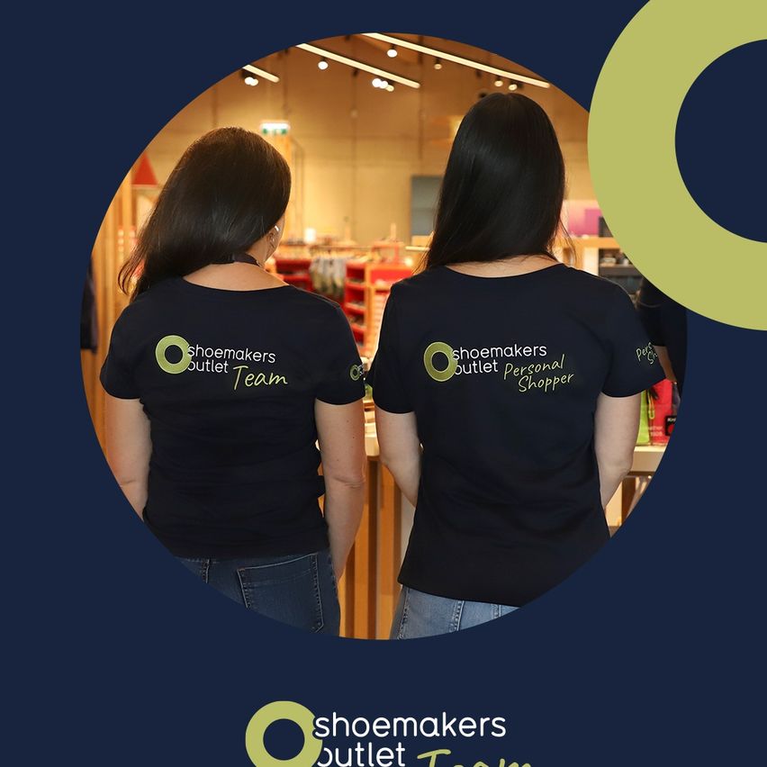 Zwei Frauen in schwarzen T-Shirts mit dem Text 'Schuhmacher Outlet Team' stehen mit dem Rücken zueinander in einem Geschäft. Der Hintergrund ist verschwommen, wobei Regale und Produkte zu sehen sind.