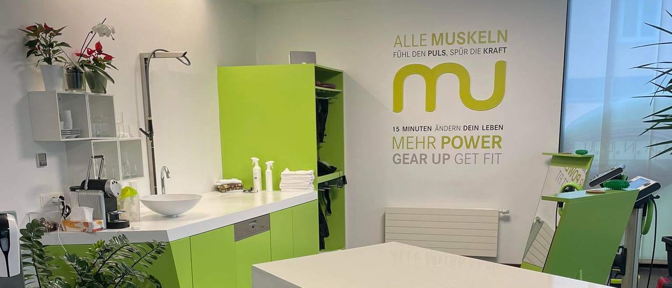 Eine Küche mit grünen Schränken und einer weißen Arbeitsplatte. Eine grüne Pflanze in einem schwarzen Topf steht auf dem Boden. Eine weiße Schüssel und ein Wasserkocher sind auf der Arbeitsplatte. An der Wand befindet sich ein gelbes und weißes Schild mit dem Wort 'mu'.