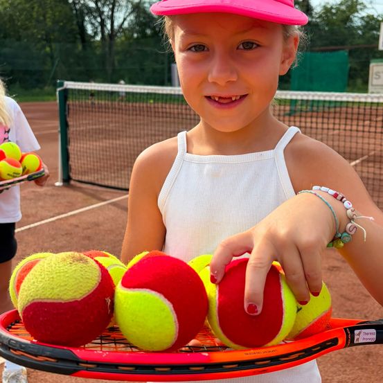 Ein junges Mädchen in einem rosa Hut hält einen Tennisschläger mit Tennisbällen darauf, lächelt und posiert für ein Foto auf einem Tennisplatz.