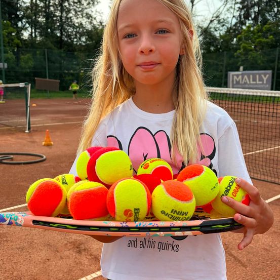 Ein junges Mädchen mit blonden Haaren hält einen Tennisschläger voller Tennisbälle und steht auf einem Tennisplatz mit einem verschwommenen Hintergrund aus Zaun und Bäumen.