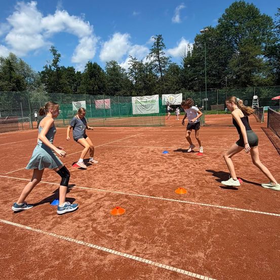 Vier Mädchen rennen auf einem Tennisplatz und tragen Turnschuhe und Knieschützer. Sie folgen einem Weg, der von orangefarbenen Hütchen markiert ist. Im Hintergrund befindet sich ein Zaun mit Bannern und Bäumen.