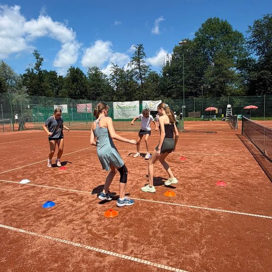 Fünf Frauen befinden sich auf einem Tennisplatz und spielen ein Spiel mit orangefarbenen und blauen Kegeln, die auf dem Boden platziert sind. Der Platz ist von einem Zaun umgeben, und es gibt Bäume und einen klaren blauen Himmel im Hintergrund.