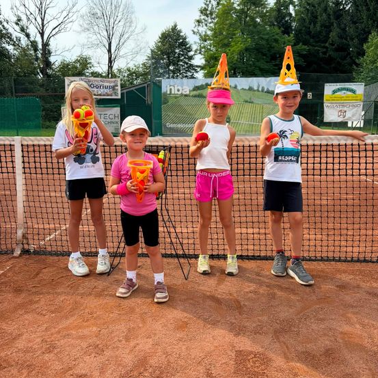 Vier Kinder stehen auf einem Tennisplatz, jedes hält einen Tennisball und trägt Kegelmützen. Sie lächeln und posieren für ein Foto.