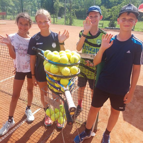 Vier Kinder stehen auf einem Tennisplatz, lächeln und posieren für ein Foto. Sie halten Tennisschläger und stehen neben einem Korb voller Tennisbälle. Im Hintergrund ist ein Tennisplatz mit Netz und einem Rasenfeld zu sehen.
