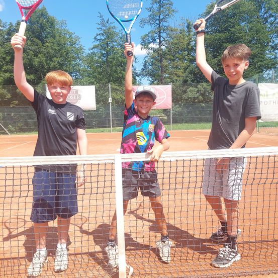 Drei junge Jungen stehen auf einem Tennisplatz und halten Tennisschläger. Sie lächeln und scheinen für ein Foto zu posieren. Der Junge in der Mitte trägt ein buntes Shirt. Hinter ihnen steht ein Zaun mit Bannern.