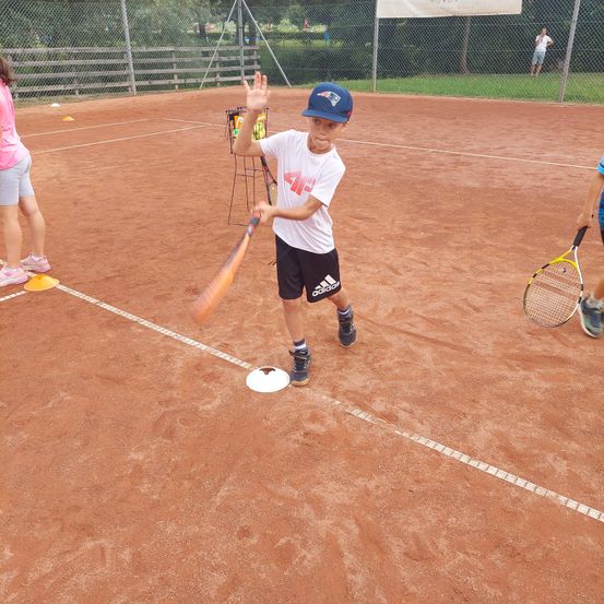 Ein junger Junge übt das Schlagen eines Tennisballs mit einem Schläger auf einem Sandplatz. Er trägt einen blauen Hut und Turnschuhe.