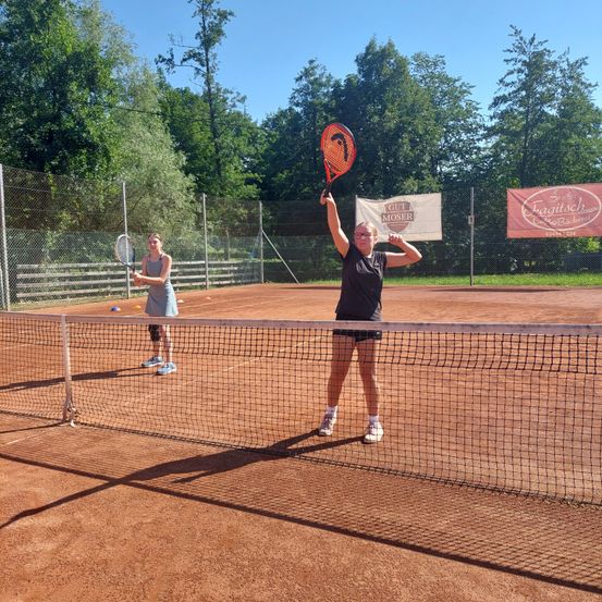 Zwei Frauen spielen Tennis auf einem Sandplatz, eine in einem schwarzen Shirt und die andere in einem blauen Kleid. Sie halten beide Tennisschläger.