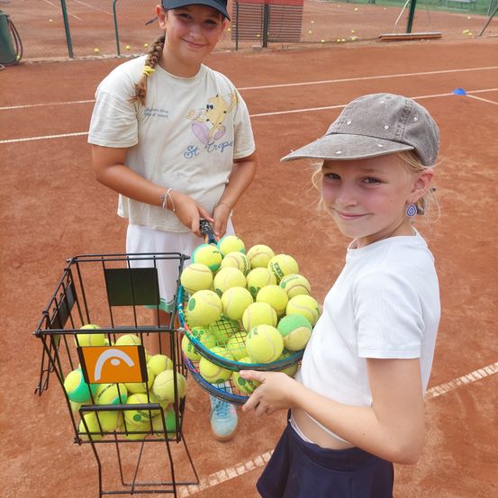 Zwei junge Mädchen stehen auf einem Tennisplatz, eine hält einen Korb mit Tennisbällen und die andere eine Tennisschläger. Sie lächeln beide und scheinen für ein Foto zu posieren.