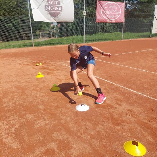 Ein junges Mädchen übt Tennis auf einem Sandplatz und beugt sich vor, um einen Tennisball in der Nähe eines gelben Kegels aufzuheben.