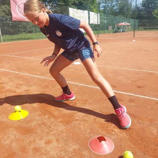 Ein junges Mädchen in einem blauen Shirt und rosa Turnschuhen übt Tennis auf einem Sandplatz, mit zwei Tennisbällen und Kegeln in der Nähe.