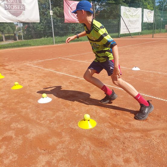 Ein junger Junge übt Tennis auf einem Sandplatz und läuft auf Tennisbälle zu, die auf dem Boden platziert sind. Er trägt ein grünes und schwarzes Shirt mit dem Wort 'Nike' darauf.