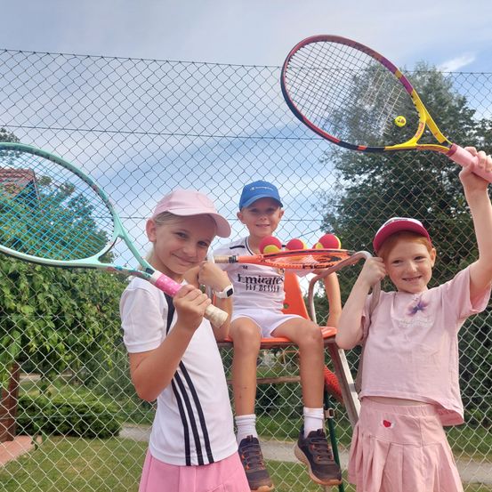 Drei Kinder, zwei Mädchen und ein Junge, posieren mit Tennisschlägern in einem Park. Das Mädchen links trägt einen rosa Hut und ein weißes Shirt. Der Junge in der Mitte sitzt auf einem Stuhl mit einem Schläger in den Händen. Das Mädchen rechts trägt einen rosa Hut und einen rosa Rock.