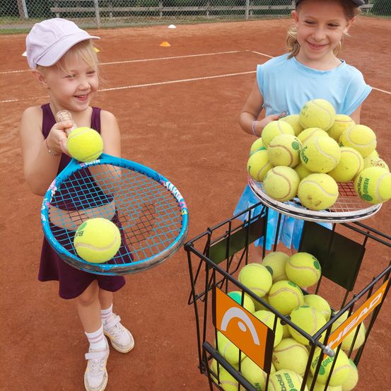 Zwei junge Mädchen auf einem Tennisplatz, eine hält einen Tennisschläger mit Tennisbällen und die andere einen Korb voller Tennisbälle.