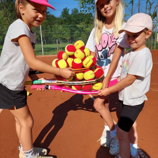 Drei junge Mädchen, die passende Mützen tragen, halten einen Korb mit Tennisbällen auf einem Tennisplatz. Sie lächeln und sehen aus, als wären sie bereit zu spielen.