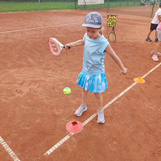 Ein kleines Mädchen übt Tennis auf einem Sandplatz, hält einen Schläger und steht in der Nähe eines Tennisballs und von Kegeln.