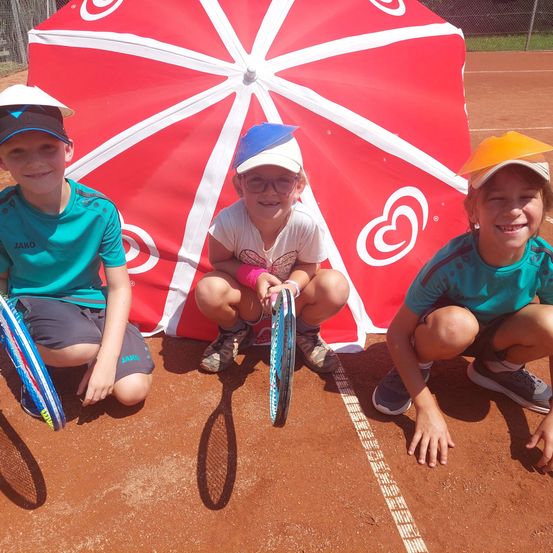 Drei junge Jungen in Tenniskleidung knien lächelnd auf einem Tennisplatz, mit einem roten Regenschirm mit weißen Herzen im Hintergrund.