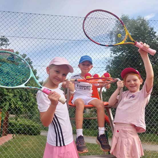 Drei Kinder, zwei Mädchen und ein Junge, posieren für ein Foto mit Tennisschlägern. Der Junge sitzt auf einem Stuhl, während die Mädchen auf beiden Seiten von ihm stehen. Sie alle lächeln und halten Tennisschläger.