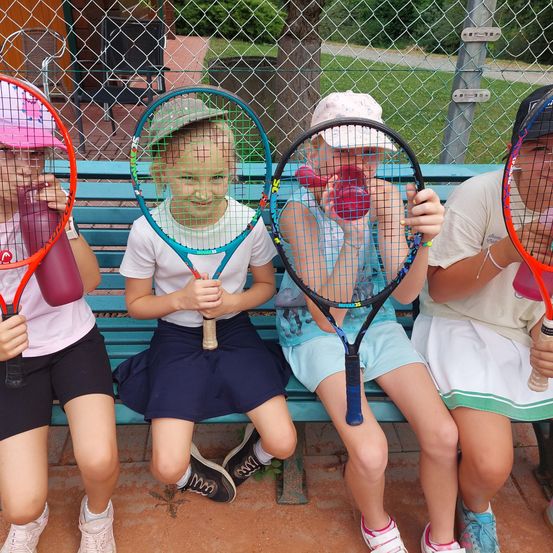Fünf Kinder sitzen auf einer Bank, halten Tennisschläger und Wasserflaschen in der Hand und lächeln für ein Foto. Dahinter trennt ein Maschendrahtzaun den Bereich von einem Grasfeld.