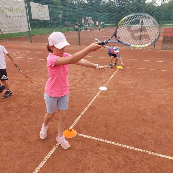 Ein junges Mädchen in einem rosa Shirt und Hut übt Tennis auf einem Sandplatz, schlägt einen Ball mit einem Schläger. Andere Spieler sind im Hintergrund, und orange Kegel markieren das Spielfeld.