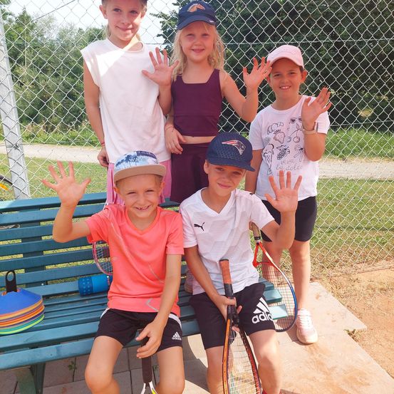 Fünf Kinder, vier Mädchen und ein Junge, posieren für ein Foto auf einem Tennisplatz. Sie alle lächeln und winken. Der Junge in der Mitte hält eine Tennisschläger.