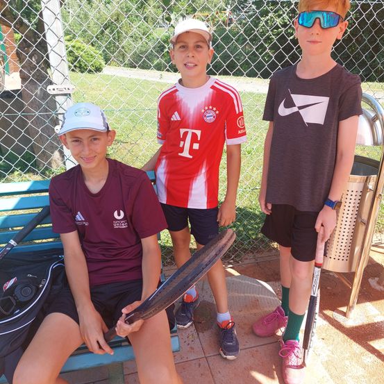 Drei Jungen in Sportkleidung, zwei stehend und einer auf einer Bank sitzend, halten einen Baseballschläger, mit einem Mülleimer und einem Maschendrahtzaun im Hintergrund.