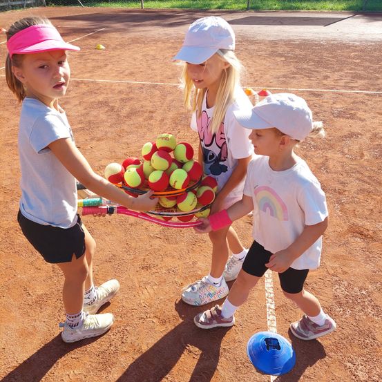 Drei junge Mädchen auf einem Tennisplatz, eine hält einen Korb voller Tennisbälle, eine andere eine Tennisschläger und die dritte einen Frisbee.